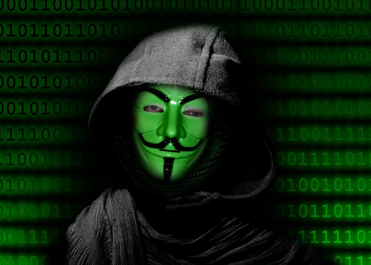 Man in Anonymous mask standing in front of a screen of computer code.