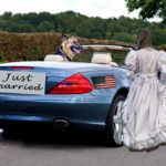 German Shepherd in driver's seat of blue convertible waiting for woman wearing wedding dress.