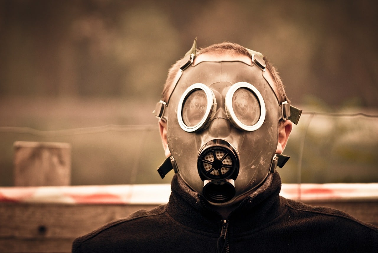Man in a gas mask and hazmat suit standing outside in a dirty field.