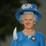 A doll of Queen Elizabeth wearing a blue dress and hat and waving.