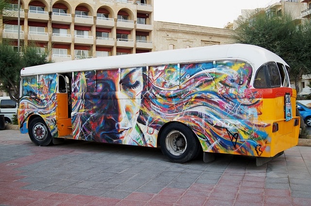 Rainbow colored school bus with a photo of a woman on the side near a building.