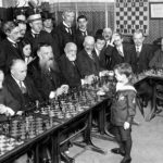 8 year old boy standing in front of a table with twenty men after he beat them in chess.