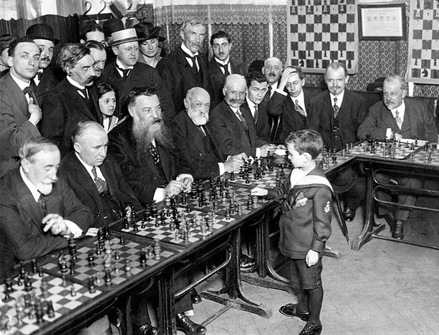 8 year old boy standing in front of a table with twenty men after he beat them in chess.