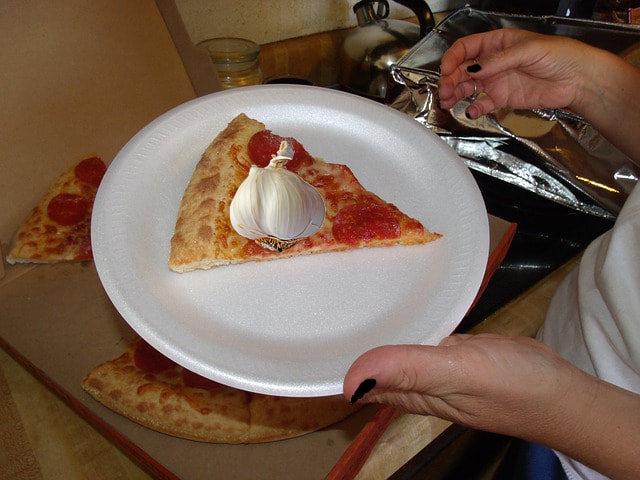 Man in kitchen holding plastic plate with slice of pizza on it.