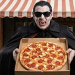 Vampire holding a pizza with a lot of garlic on it in front of a Italian restaurant.