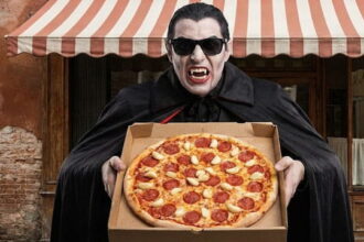 Vampire holding a pizza with a lot of garlic on it in front of a Italian restaurant.