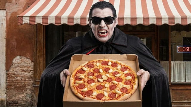 Vampire holding a pizza with a lot of garlic on it in front of a Italian restaurant.