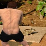 Shirtless man bending over to talk to a chipmunk in forest.