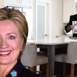 Bill Clinton in black suit and Hillary Clinton in black suit standing inside white kitchen.
