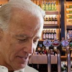 Joe Biden standing inside bar surrounded by alcohol bottles.