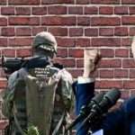Joe Biden wearing blue suit near Army soldier in front of brick wall.
