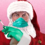 Santa Claus wearing blue face mask while holding a long medical syringe filled with coronavirus vaccine.