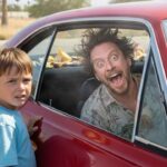 Young son standing in front of sports car in large parking lot with his dad inside of the car.