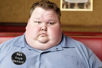 Fat obese man in gray shirt standing in front of buffet table.