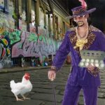 Pimp dressed in purple suit surrounded by chickens in dark alley.