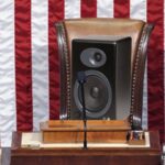 Music speaker sitting on Congress House Speaker chair in front of American flag.