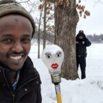 Somali man standing next to a lightbulb taped to a broomstick with an ICE agent in the background of Minnesota.