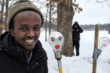 Somali man standing next to a lightbulb taped to a broomstick with an ICE agent in the background of Minnesota.