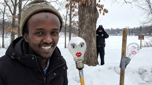 Somali man standing next to a lightbulb taped to a broomstick with an ICE agent in the background of Minnesota.