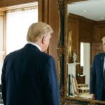 Donald Trump staring into a mirror in a large room.