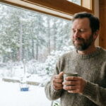Unvaccinated American man holding a mug of tea looking out house window at snow.