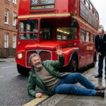 British man lying on the ground on the street in front of a big red bus in England, UK.
