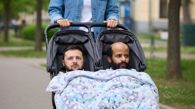 Woman pushing a baby stroller with Zelensky and a Somali man in it in front if an IRS tax building.