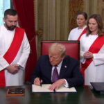 Donald Trump wearing a suit sitting behind a desk with five doctors wearing white robes and red sashes standing behind him.