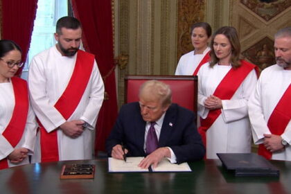 Donald Trump wearing a suit sitting behind a desk with five doctors wearing white robes and red sashes standing behind him.