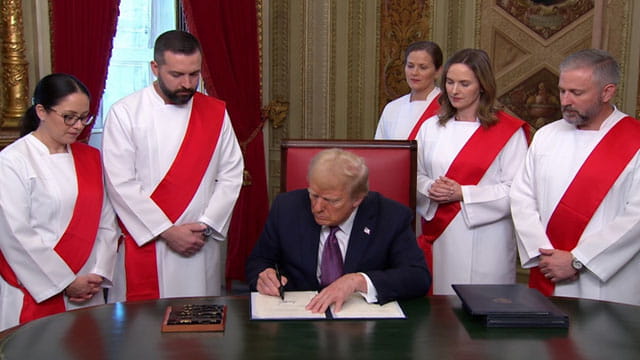 Donald Trump wearing a suit sitting behind a desk with five doctors wearing white robes and red sashes standing behind him.