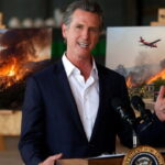 California Governor Gavin Newsom speaking at a podium in front of images of wildfires.