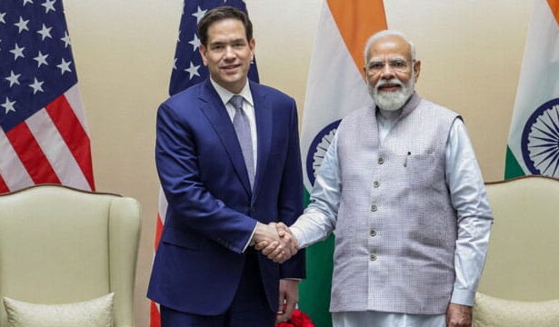 Marco Rubio shaking hands with Indian Prime Minister Modi on stage in front of flags.