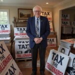 Bernie Sanders standing in a home next to Tax the Trillionaire signs with Elon Musk's face on them.