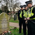 UK police officers standing in front of a tombstone of a deceased white British man inside of a graveyard.