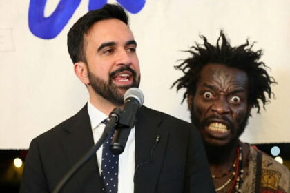 NYC Mayor Zohran Mamdani giving a speech on stage in front of a Haitian migrant.