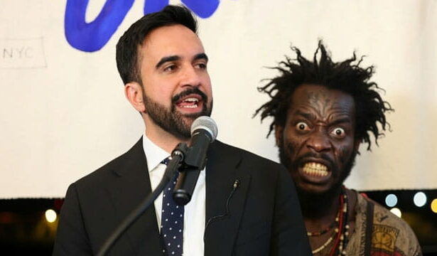 NYC Mayor Zohran Mamdani giving a speech on stage in front of a Haitian migrant.