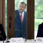 Donald Trump and Mark Carney sitting at a table talking while Ted Cruz stands outside of a window looking inside.