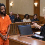 Black criminal wearing orange jumpsuit in court in front of white female judge wearing black robe.