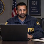 Indian FBI Director Director Kash Patel wearing an FBI jacket sitting at desk staring at a laptop.