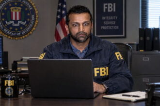 Indian FBI Director Director Kash Patel wearing an FBI jacket sitting at desk staring at a laptop.