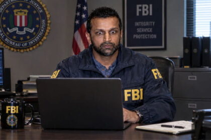 Indian FBI Director Director Kash Patel wearing an FBI jacket sitting at desk staring at a laptop.