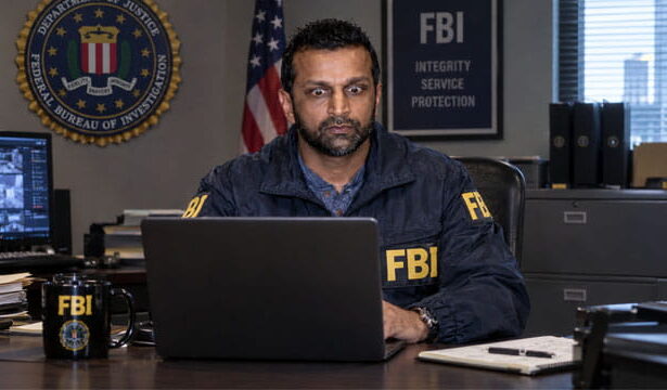 Indian FBI Director Director Kash Patel wearing an FBI jacket sitting at desk staring at a laptop.