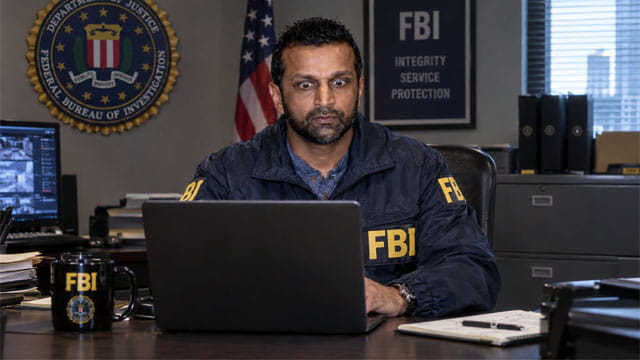 Indian FBI Director Director Kash Patel wearing an FBI jacket sitting at desk staring at a laptop.