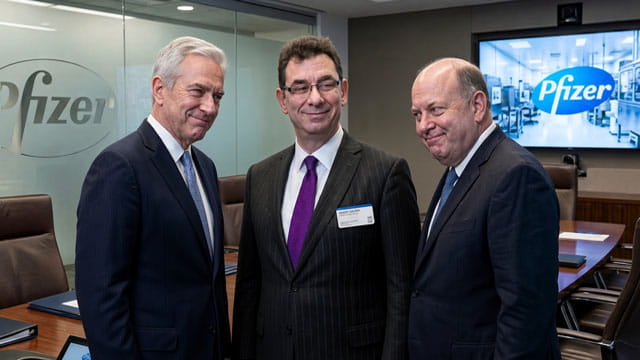 Pfizer CEO man in suit standing in the middle of two other suited executives in a boardroom.