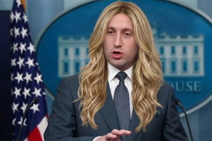 Trump Chief of Staff Stephen Miller wearing a long blonde wig speaking behind a White House Press Secretary podium.